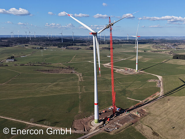 Windenergieanlagen auf einem grünen Feld. Im Vordergrund steht eine Windenergieanlage mit einem roten Baukran.
