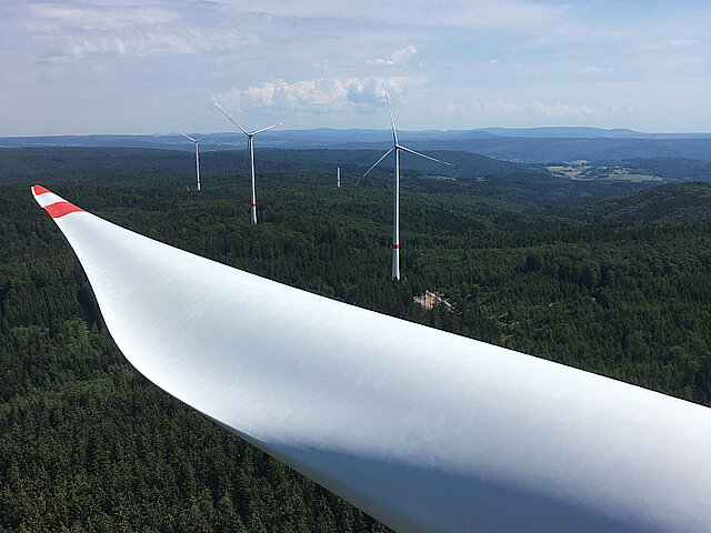 Zu sehen ist das Rotorblatt einer Windenergie-Anlage. Das Foto wurde auf der Turbine der Anlage aufgenommen. Im Hintergrund sind weitere Windenergie-Anlagen im Wald zu sehen.