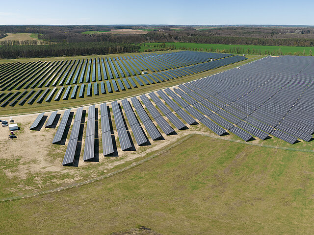 Ein Bild des Solarparks aus der Vogelperspektive. Im Hintergrund sind Hügel erkennbar, der Solarpark wird von niedrigen Bäumen und Stäuchern umsäumt. 