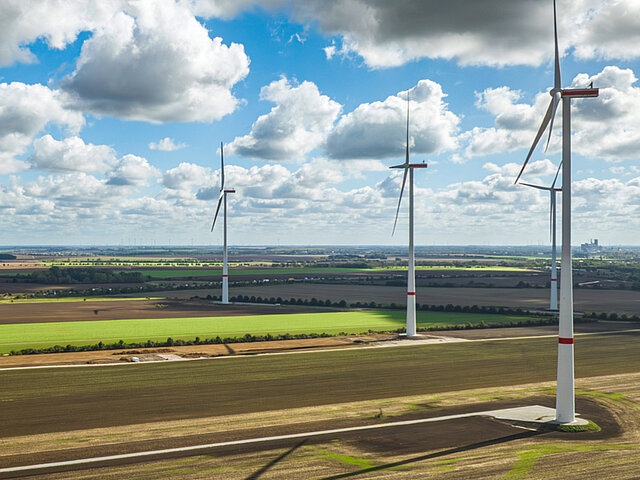 Drei Windräder stehen auf einem Feld. Die Sonne scheint, es sind einige Wolken am Himmel.