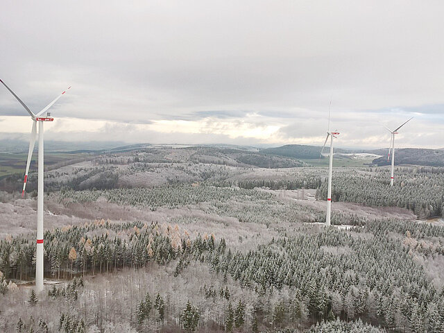 Die drei Anlagen des Windparks ragen in die Landschaft vor der winterlichen Szenerie verschwimmen sie mit dem grau des Himmels und den schneebedeckten Bäume. Die Schneedecke reicht bis zu den Hügeln am Horizont.