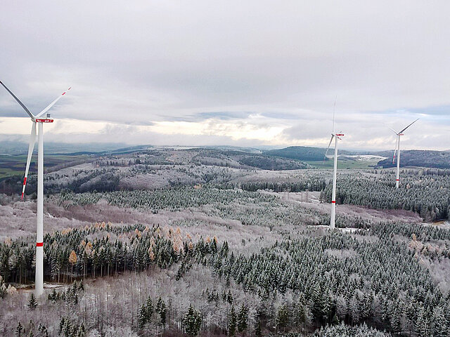 Die drei Anlagen des Windparks ragen in die Landschaft vor der winterlichen Szenerie verschwimmen sie mit dem grau des Himmels und den schneebedeckten Bäume. Die Schneedecke reicht bis zu den Hügeln am Horizont.