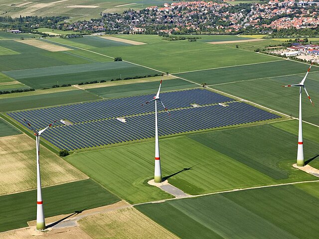Auf einer landwirtschaftlich genutzten Fläche stehen drei WEA, dahinter ist ein Solarpark zu sehen. Am Horizont liegt eine Gemeinde.