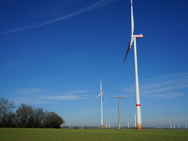 Zu sehen sind mehrere Windenergie-Anlagen des Windparks Biebelnheim-Gabsheim. Sie ragen in den bleuen Himmel. Im Vordergrund verläuft eine Stromtrasse.