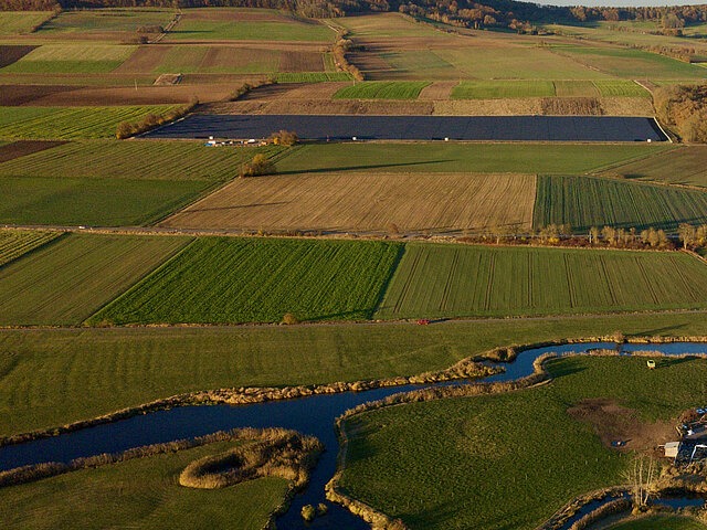 Im Hintergrund ist der Solarpark Röckingen zu sehen. Im Vordergrund steht ein Flusslauf und mehrere Äcker.