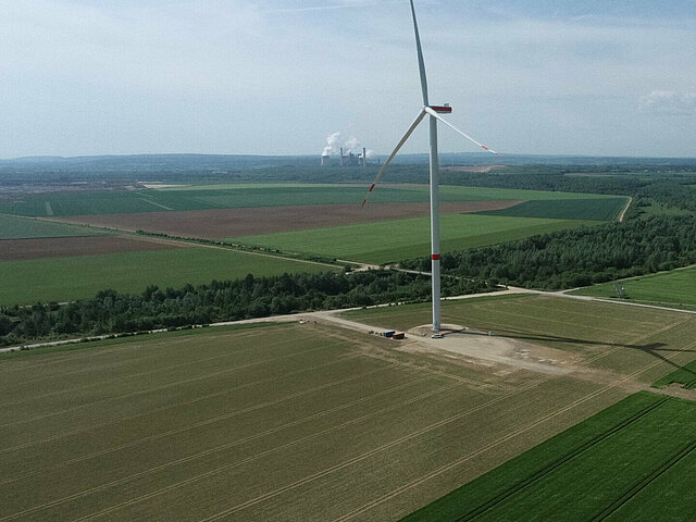 Eine Windenergie-Anlage steht auf einem Acker. IM Hintergrund sind die rauchenden Türme eines Industriebetrieb zu sehen.