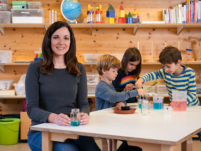 Eine Erzieherin sitzt mit experimentierenden Kindern am Tisch.