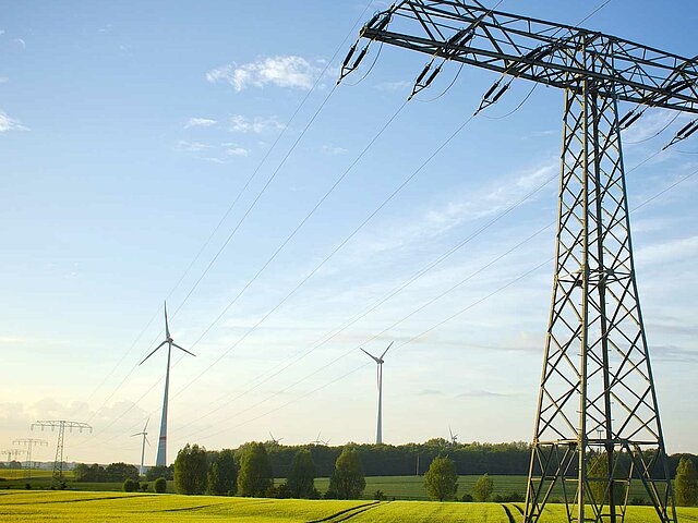 Im Vordergrund steht ein großer Strommast. Dahinter sind auf blühenden Rapsfeldern verschiedene Windenergie-Anlagen zu sehen.