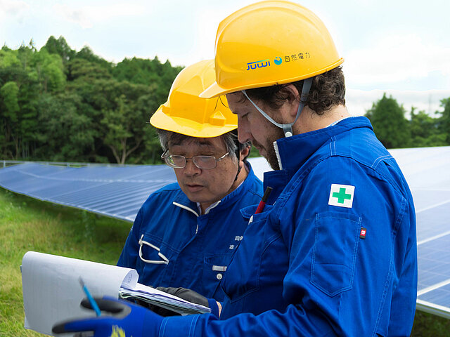 Two JUWI engineers in helmets and work attire are standing in front of solar panels and discussing.