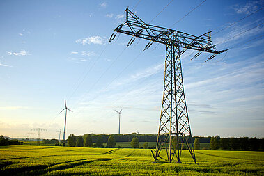 Im Vordergrund steht ein großer Strommast. Dahinter sind auf blühenden Rapsfeldern verschiedene Windenergie-Anlagen zu sehen. 