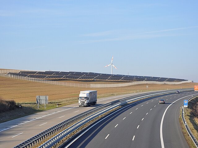 Ein Blick über eine dreispurige Autobahn. Am Horizont ist eine große PV-Anlage und mehrere Windräder zu sehen.