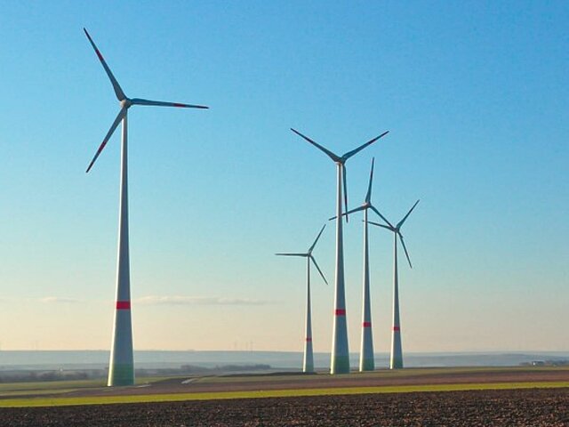 Windräder auf einem Feld, Acker im Vordergrund und blauer Himmel im Hintergrund.