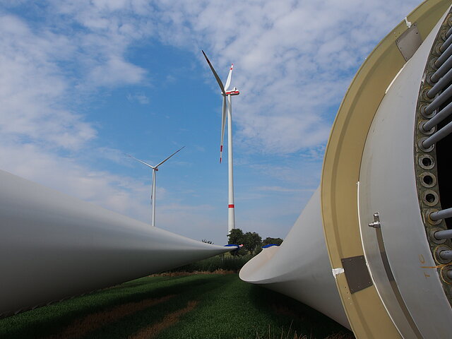 Zwei Rotorblattflügel einer Windenergie-Anlage liegen auf dem Boden und warten darauf montiert zu werden. Im Hintergrund sind zwei bereits fertiggestellte Windenergie-Anlagen zu sehen.