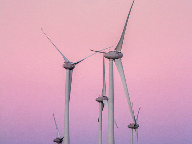 Mehrere Windenergie-Anlagen ragen in einen leuchtenden rosa Himmel. 