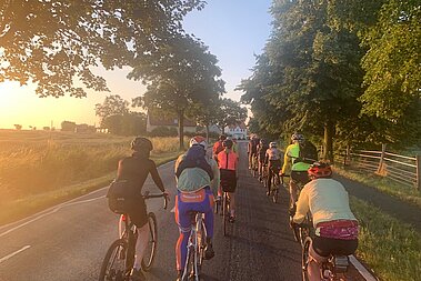 Eine Gruppe Radler*innen sitzen auf ihren Rennrädern und fahren auf einer Allee.