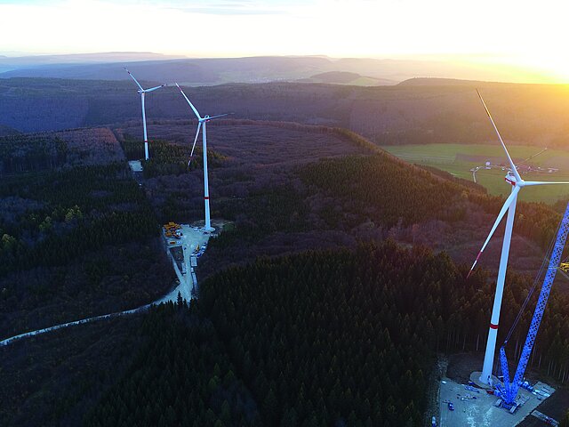 Drei Windenergie-Anlagen in einer forstwirtschaftliche genutzten Gebiet aus der Vogelperspektive. Im Morgengrauen sind die ersten Sonnenstraheln zu sehen. An der vordersten der Anlagen steht ein großer blauer Baukran.