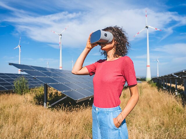 Frau steht mit VR-Brille vor einem Solarpark und Windenergieanlagen.