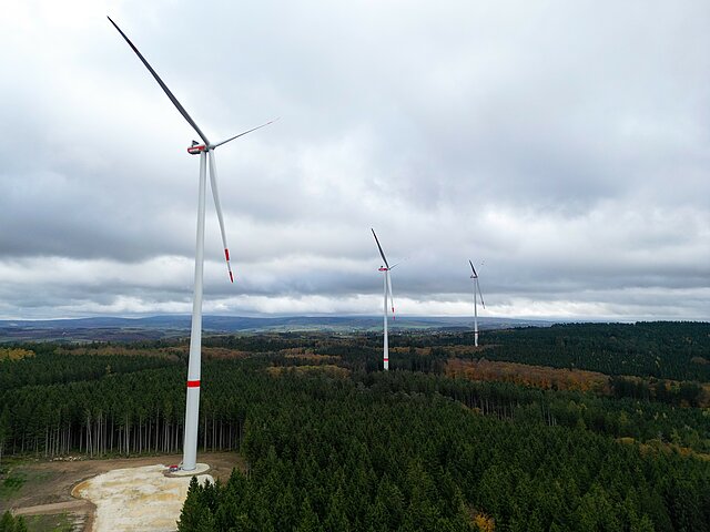 Drei Windenergie-Anlagen des Windparks Bescheid-Süd stehen auf forstwirtschaftlich genutzten Flächen. Der Himmel ist bewölkt. Am Horizont ist klein eine Gemeinde zu sehen.