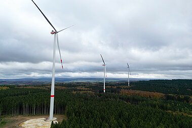 Drei Windenergie-Anlagen des Windparks Bescheid-Süd stehen auf forstwirtschaftlich genutzten Flächen. Der Himmel ist bewölkt. Am Horizont ist klein eine Gemeinde zu sehen.