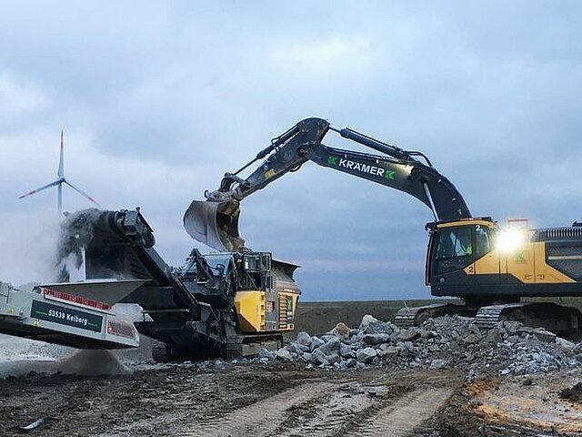Ein Bagger schaufelt den Schutt der alten Fundamente in einen Zerkleinerer. Der Himmel ist grau.