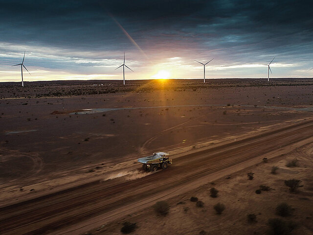 A mining truck is driving on a lonely road. On the horizon, four wind turbines can be seen.