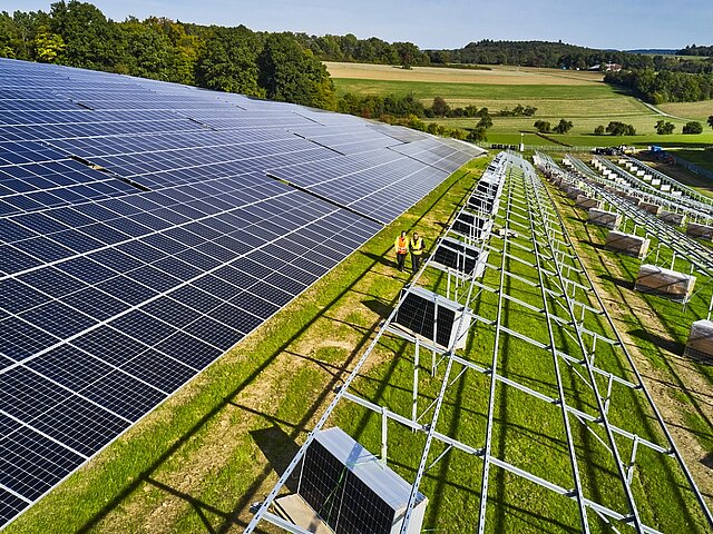 Auf der linken Seite steht ein augenscheinlich fertiger Solarpark, auf der rechten Seite stehen lediglich die Modulständer. Dazwischen laufen zwei Mitarbeiter über die Baustelle.