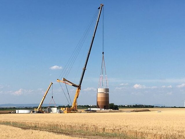 Der Betontrum wird montiert. Der Baukran hebt einen der Ringe auf den Turm, wo dieser im Anschluss befestigt wird. Im Vordergrund des Bildes steht ein Weizenacker, der Kran ragt in den blauen Himmel.
