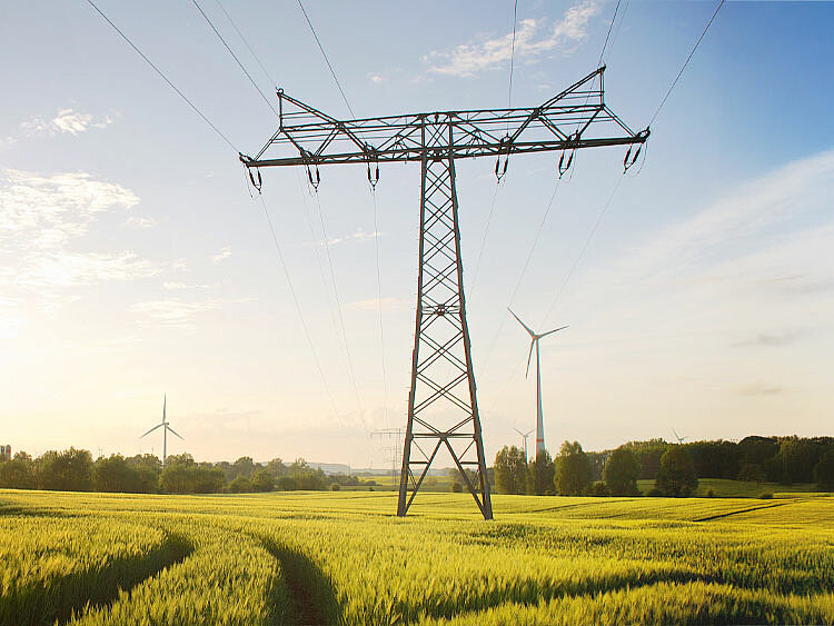 Ein Strommast steht auf einem Ackerm am Horizont ist eine Windenergie-Anlage erkennbar.