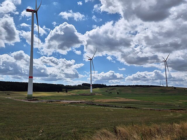 Drei Windenergieanlagen sind auf einem Feld zu sehen vor einem blauen Himmel mit vielen Wolken.