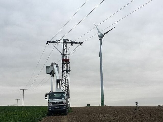 Eine Hebebühne wird ausgefahren, um Arbeiter an der Stromtrasse erste bauvorbereitenden Maßnahmen zu ergreifen. Der Himmel ist grau. Im Hintergrund ist eine der alten Anlagen zu sehen.