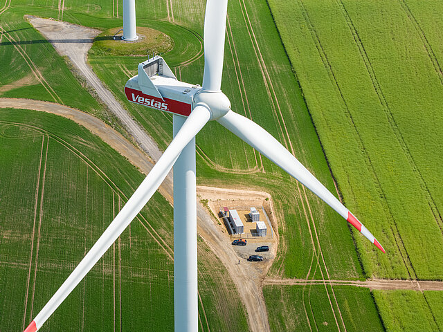 Windenergieanlage aus der Vogelperspektive. Am Boden sind landwirtschaftlich genutzte Flächen zu sehen, die grün in der Sonne erstrahlen.