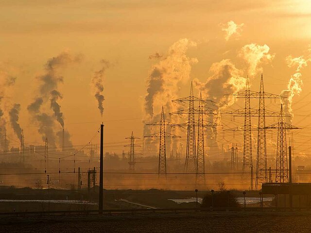 Viele rauchende Schornsteine von Industrie-Anlagen. Davor stehen jede Menge Strommasten mit Überlandkabeln.