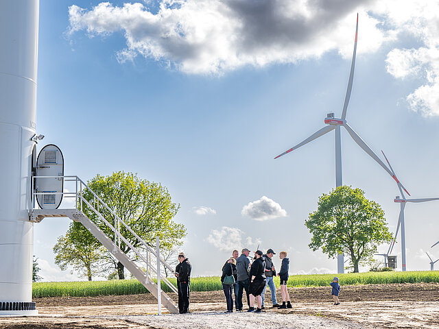 Mehrere Personen stehen or einer Windenergie-Anlage. Im Hintergrund stehen Windräder und grüne Bäume, die Sonne scheint, der Himmel ist blau mit leichten Schleierwolken.