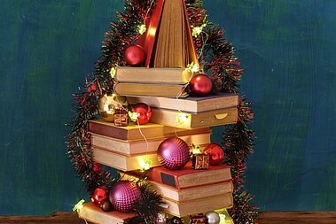 Ein Bücherstapel in Form eines Weihnachtsbaums, geschmückt mit Lichtern und Kugeln. 