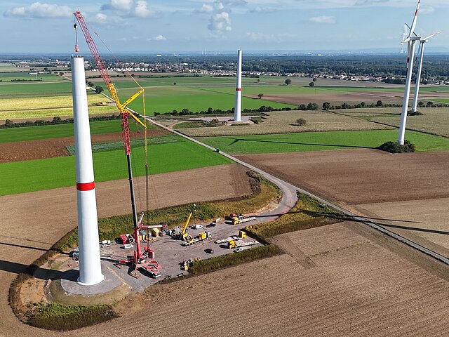 Der Turm einer Windenergieanlage wird auf einem braunen Feld aufgebaut. Umliegend gibt es weitere Felder und Windenergieanlagen.