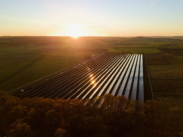 Sonnenuntergang, der Himmel ist klar und glüht am Horizont orange. Die Sonne reflektiert and den Solarmodulen. Der Solarpark schmiegt sich in die hügelige agrarwirtschaftlich genutzte Landschaft. 