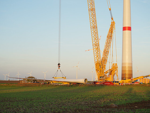 Baustelle der Windkraftanlage. Ein Baukran hebt das Rotorblatt der neuen Anlage an. Es ist früher Morgen und die Sonne taucht alles in goldenes Licht.