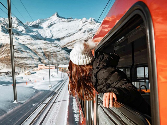 Eine junge Frau schaut aus dem Fenster eines fahrenden Zugs, im Hintergrund sind schneebedeckte Berge zu sehen.