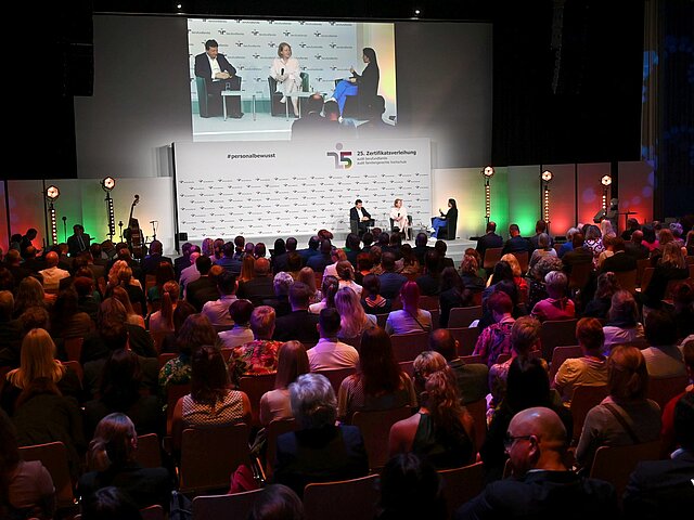 Ein Saal voller Menschen, am Kopfende sitzen Menschen auf einem Podium und diskutieren miteinander.