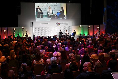Ein Saal voller Menschen, am Kopfende sitzen Menschen auf einem Podium und diskutieren miteinander. 