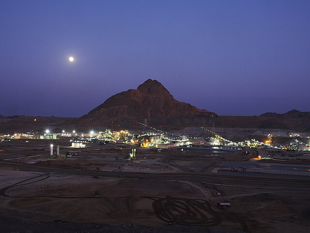 Die Mine bei Nacht. Der Mond steht über Szenerie, im Hintergrund ragt ein einzelner Berg in die Höhe. An seinem Fuß erhellen eine Vielzahl von Lichter und Industrie-Anlagen die Umgebung.