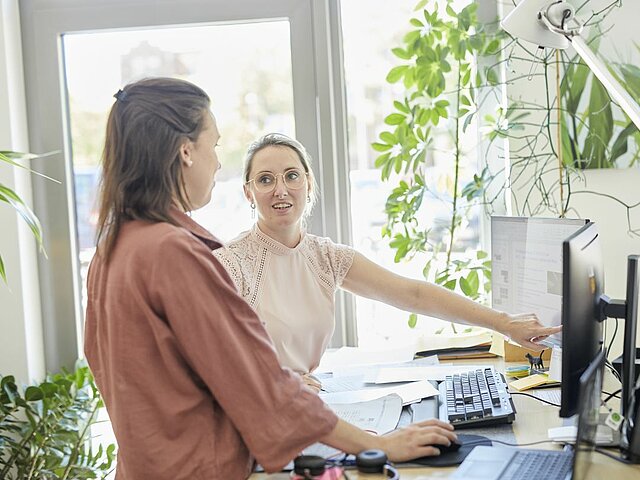 Zwei Frauen stehen im Büro vor einem Doppelbildschirm. Die eine Mitarbeiterin zeigt der anderen etwas auf dem Bildschirm. Auf dem Schreibtisch liegen Dokumente und eine Tastatur.