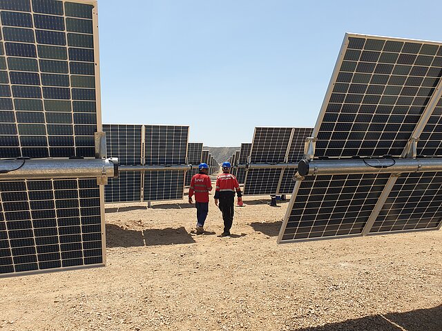 2 JUWI-Mitarbeiter arbeiten bei einem Kontrollgang zwischen den Solar-Modulen. Die Sonne scheint und der Himmel ist wolkenlos.