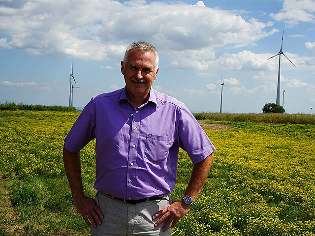 Klaus Fischer steht auf einem Feld, im Hintergrund sind Windräder zu sehen.