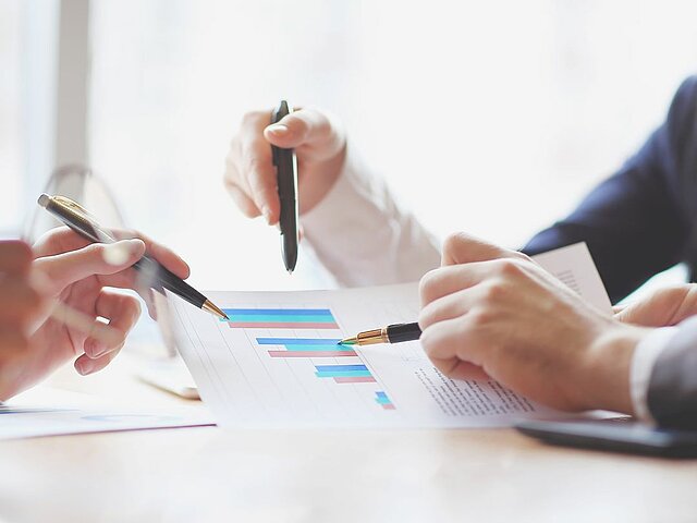 In der Mitte des Bildes ist eine Dokument mit einer Balkengrafik zu sehen. Verschiedene Personen, von denen nur die Hände und Arme zu sehen sind, schauen gemeinsam auf das Dokument. Jeder der Personen hält einen Stift in der Hand, um auf das Dokument zu zeigen und sich abzustimmen.
