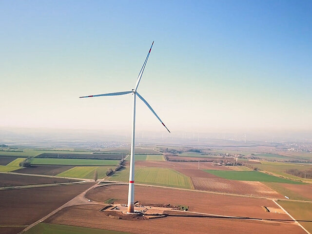 Eine einzelne Windkraft-Anlage steht auf landwirtschaftlich genutzter Fläche. Der Horizont verschwnidet im Dunst, die Sonne scheint auf die flache Landschaft ringsum.