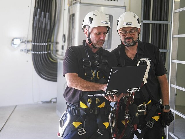 Zwei Servicetechniker stehen im Turmfuß einer Windenergieanlage und schauen zusammen in einen Laptop. Sie tragen Helme mit dem JUWI-Logo. Im Hintergrund sieht man eine Leiter und Kabel.