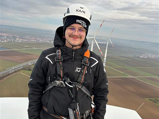 Ein Auszubildender lächelt in die Kamera, während er auf einer Windenergie-Anlage arbeitet. Im Hintergrund sind weitere Windenergie-Anlagen zu sehen.