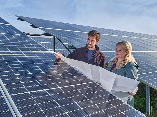 Zwei JUWI-Mitarbeiter stehen in einem Solarpark, halten einen großen Plan in der Hand und reden miteinander.