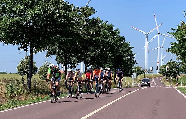 Eine Kolonne Radfahrer*innen auf einer Landstraße. Im Hintergrund sind Windräder zu sehen.